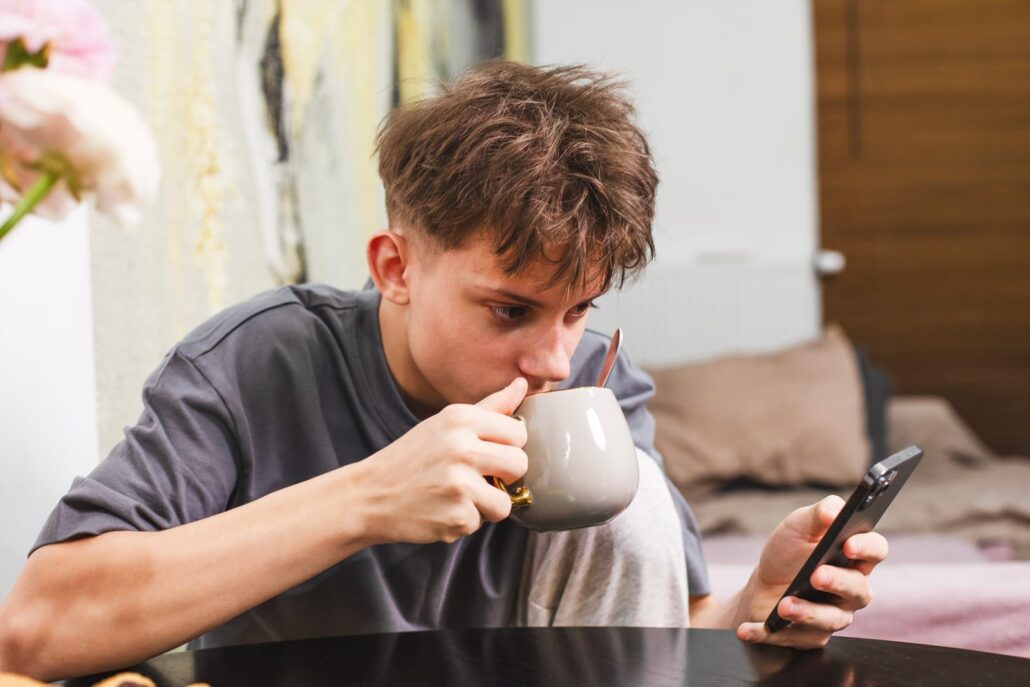 101802316 teenager enjoying tea or coffee with poppy seed cake while using smartphone in cozy kitchen digital lifestyle freelance concept