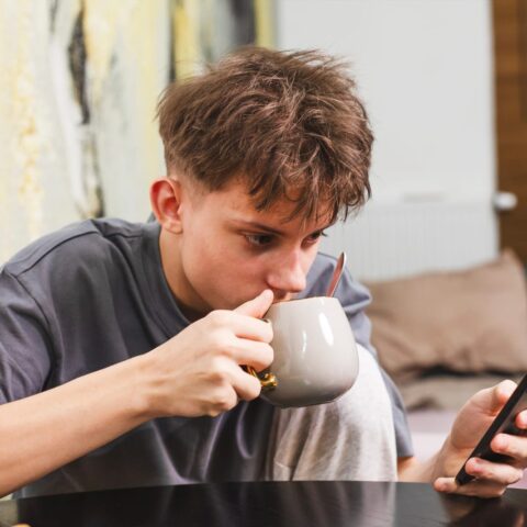 101802316 teenager enjoying tea or coffee with poppy seed cake while using smartphone in cozy kitchen digital lifestyle freelance concept